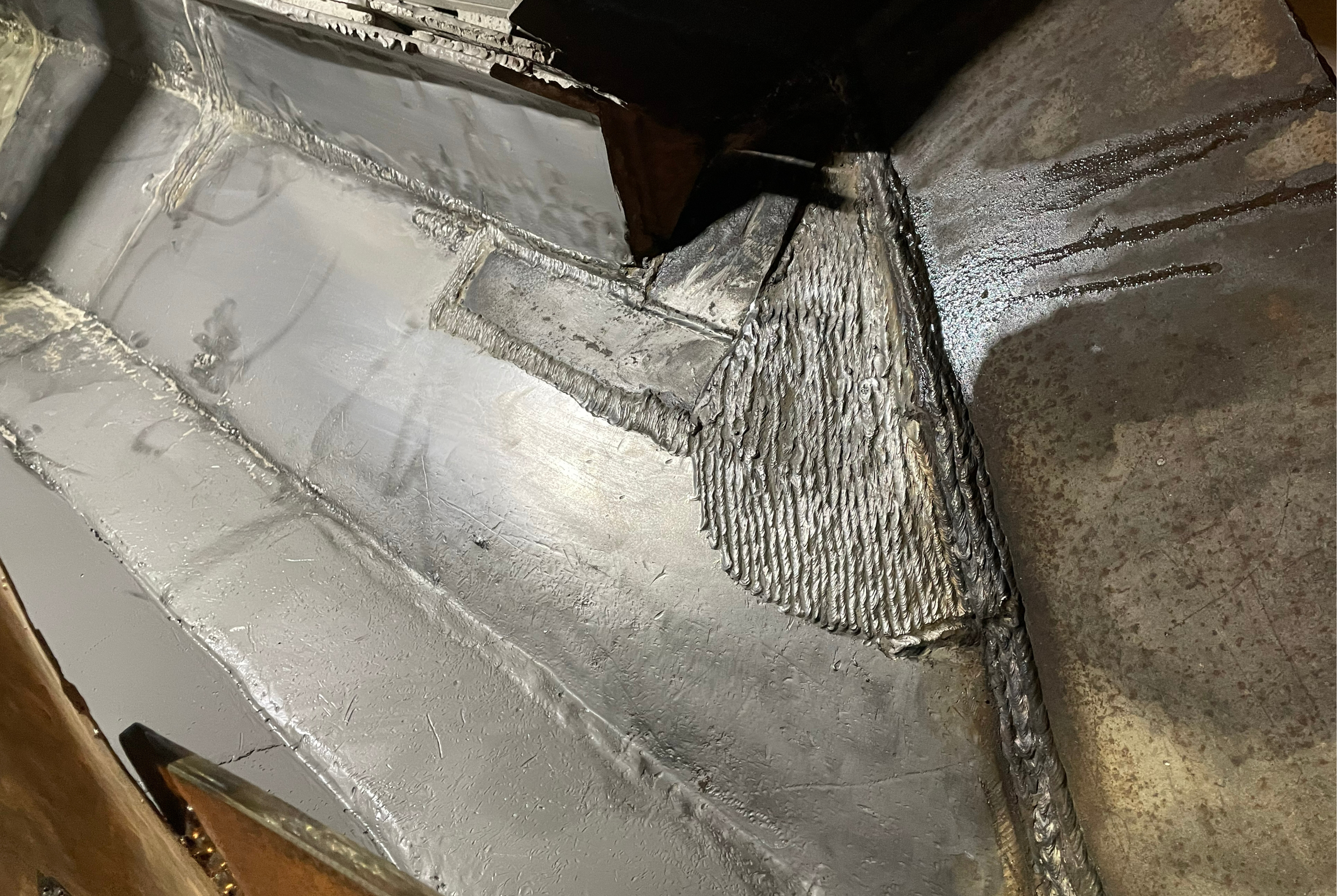 Detailed close-up view of an interior steel component structure showing a repaired area with a textured, welded patch and a grey protective coating applied across the adjacent metal joints.