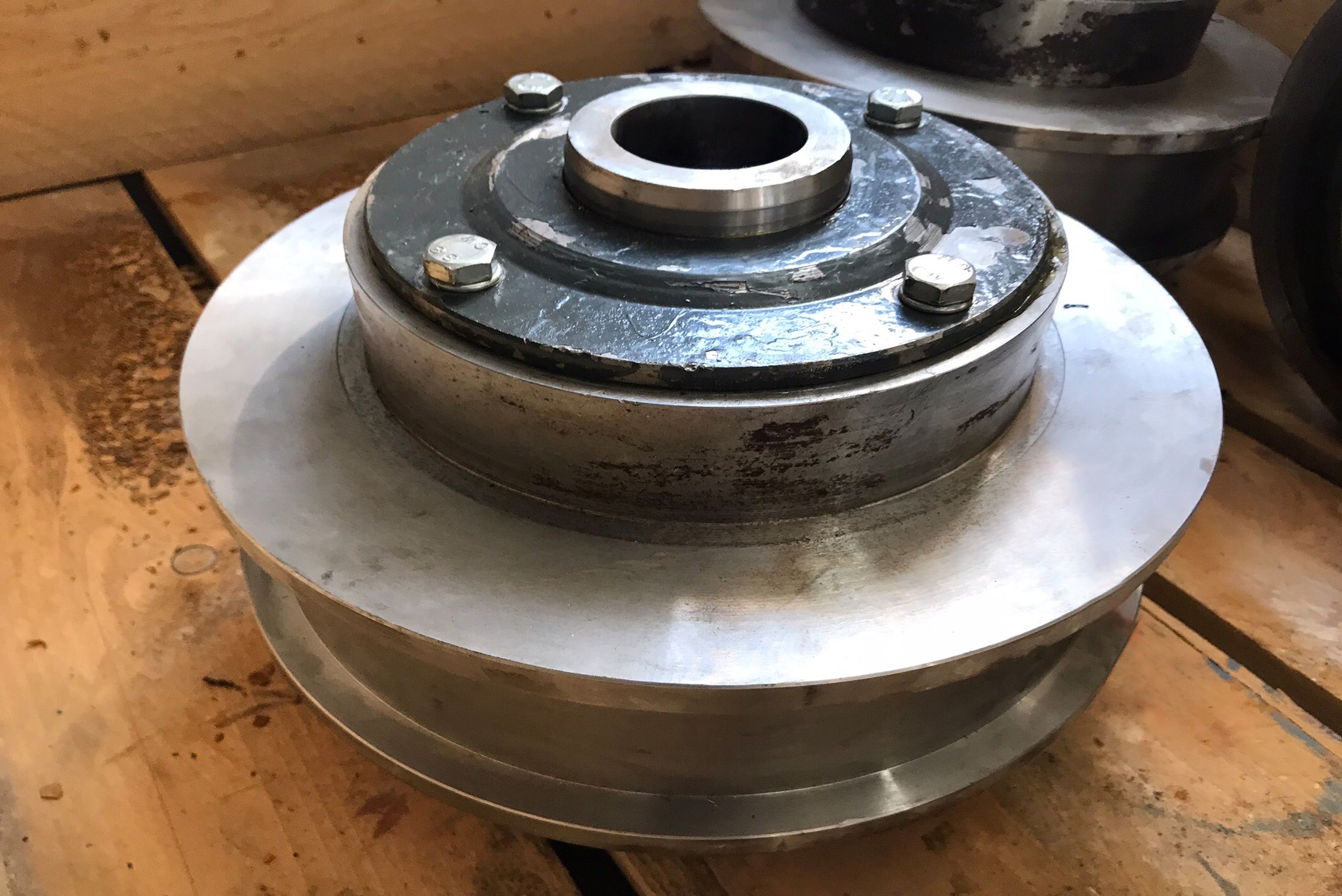 Close-up view of a circular, machined metallic pulley or flange assembly with four visible top bolts around a central bore, resting on a wooden pallet.