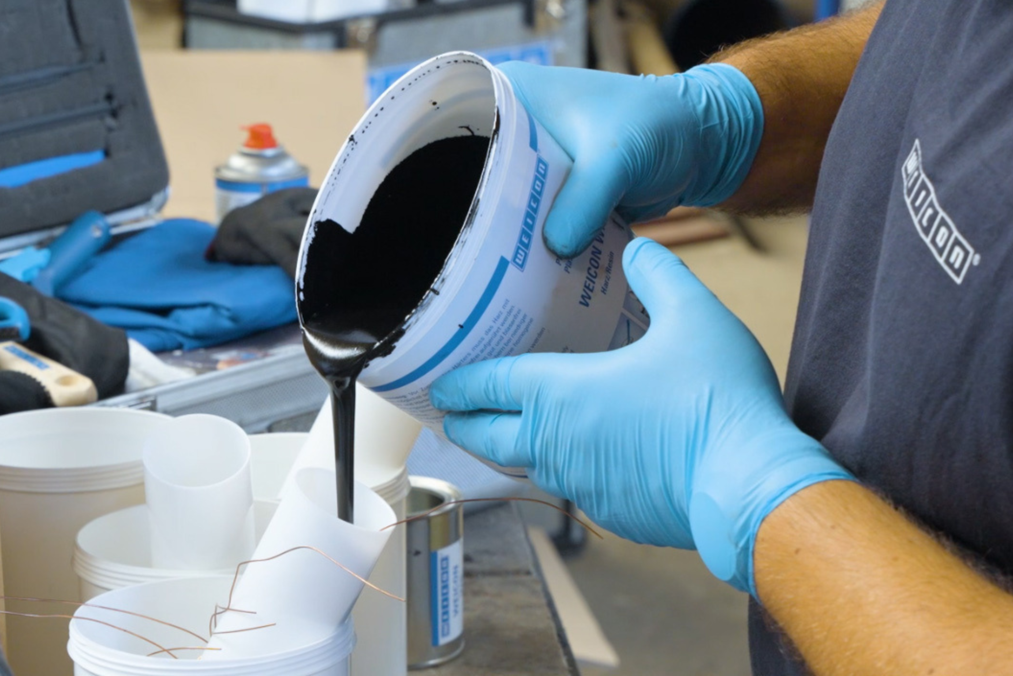 Hands wearing blue protective gloves pouring a viscous black chemical compound from a mixing bucket into a container.
