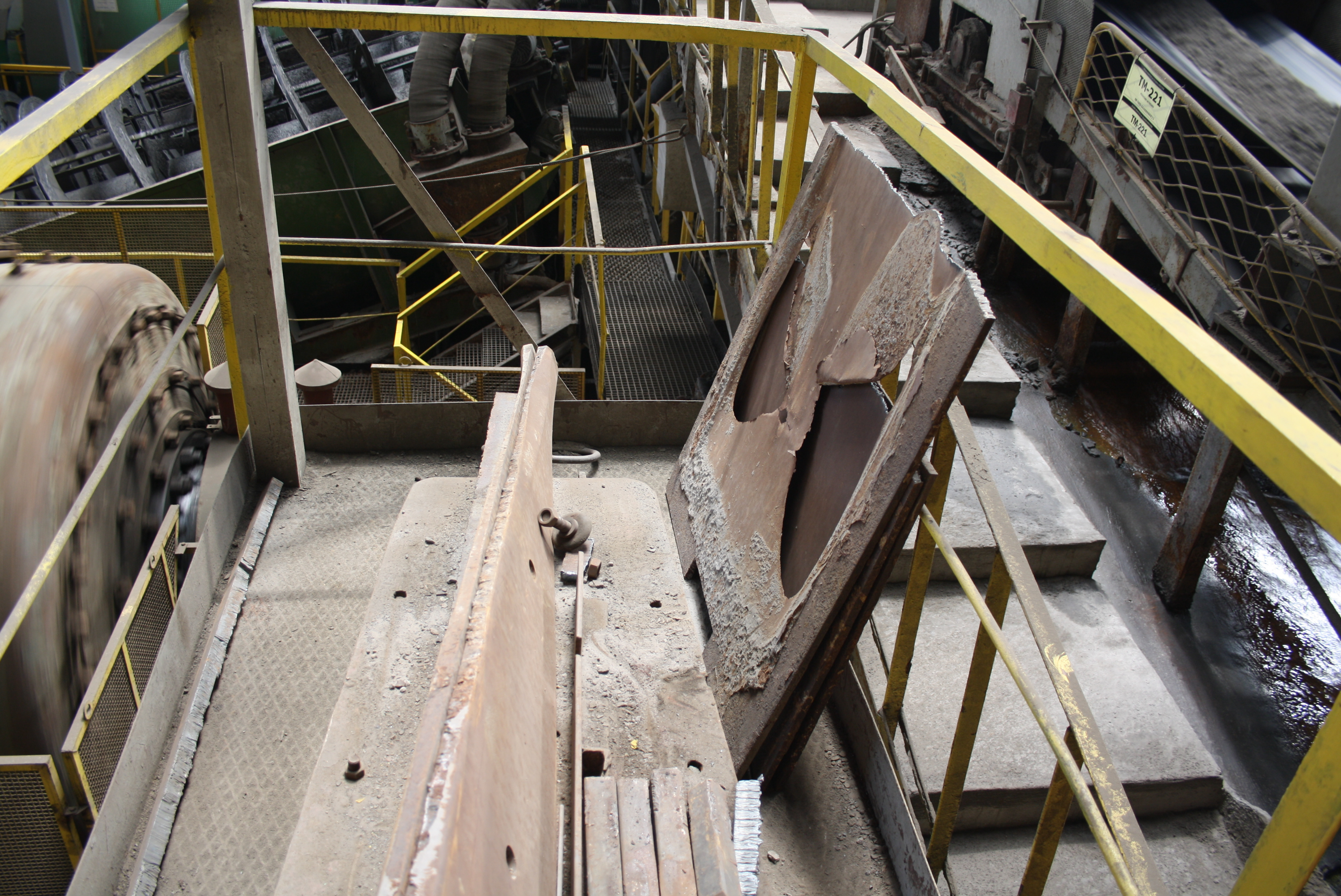 High-angle view of an industrial processing facility platform featuring worn steel plates and structural components stored near yellow safety railings, with rotating machinery and conveyor belts visible in the surrounding area.