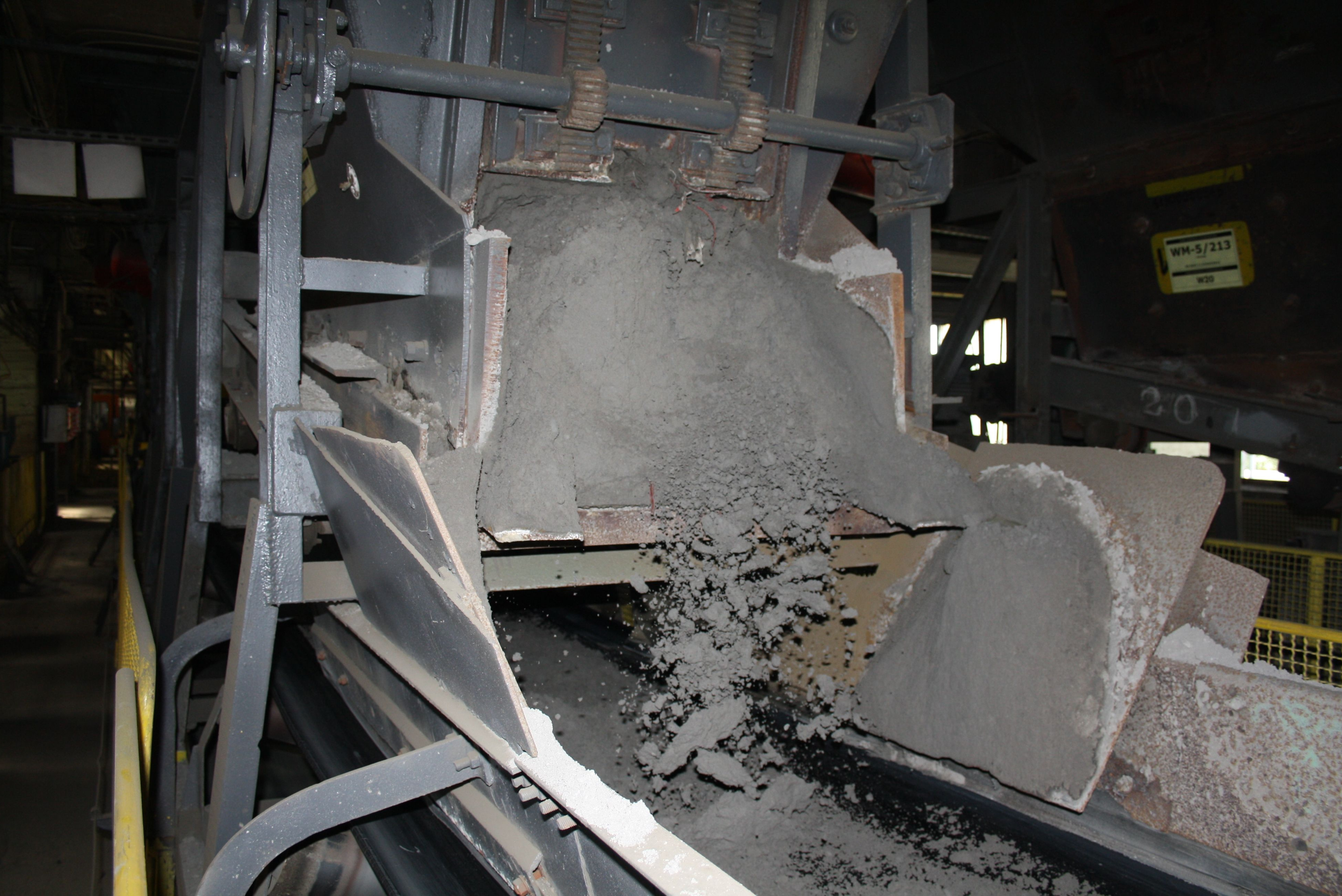 Industrial conveyor belt system discharging fine grey bulk material from a chute onto a moving conveyor belt inside a processing facility.