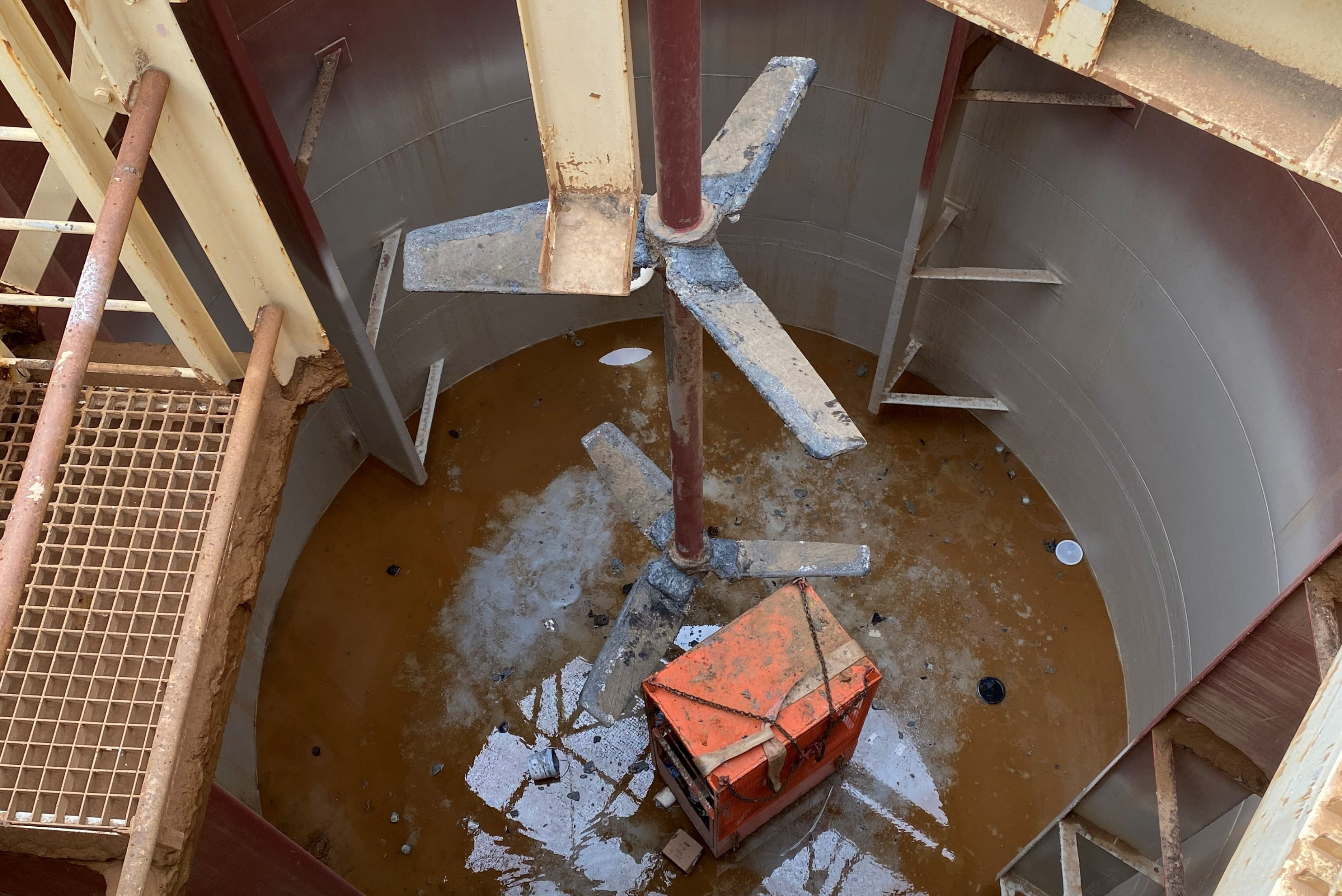 A top-down view looking into a large cylindrical industrial mixing tank, featuring a central vertical agitator shaft with multi-blade impellers and an orange equipment box positioned at the bottom