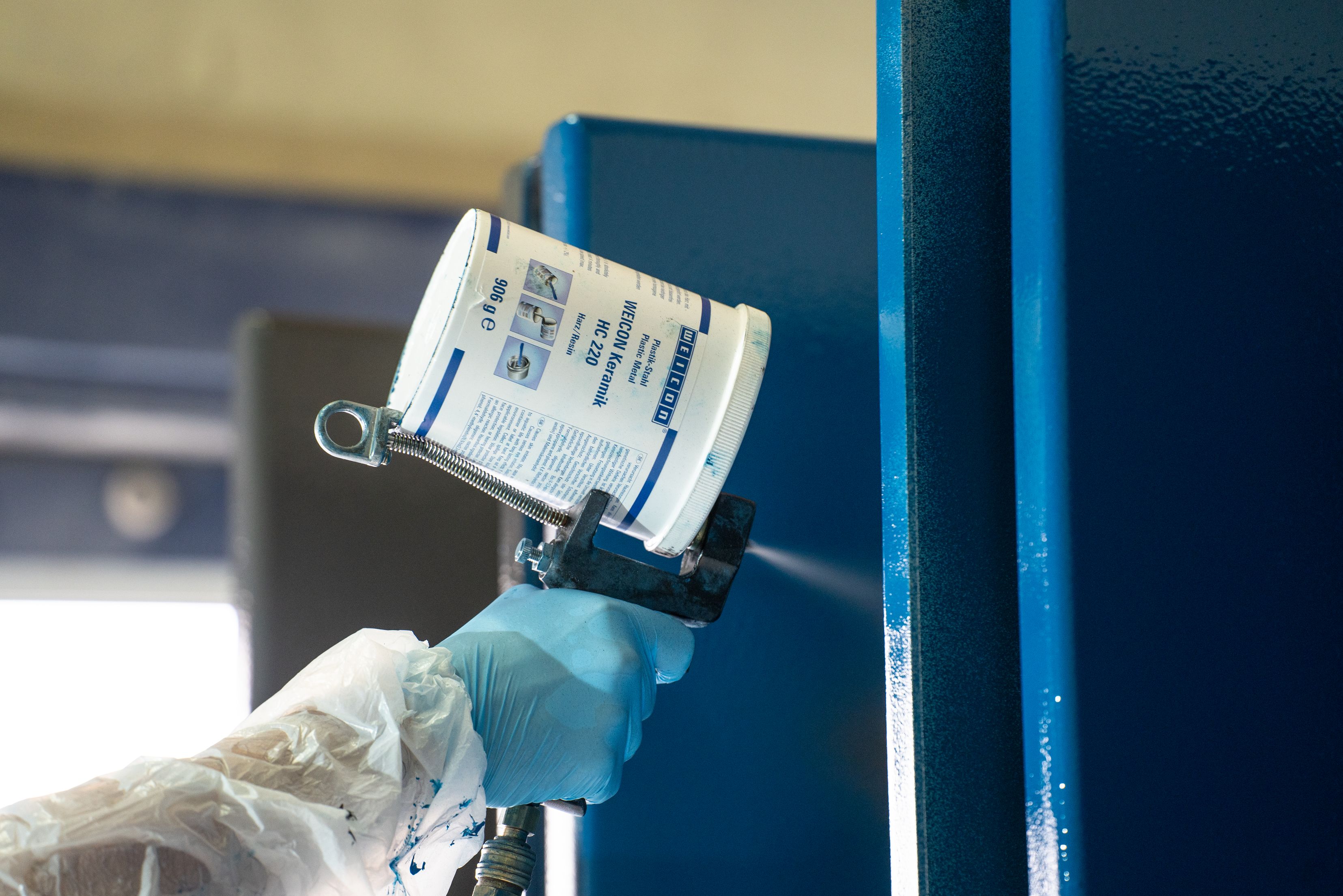 Close-up view of an operator wearing protective clothing and nitrile gloves using a spray gun fitted with a top-mounted material canister to apply a blue protective coating onto a vertical metal surface.