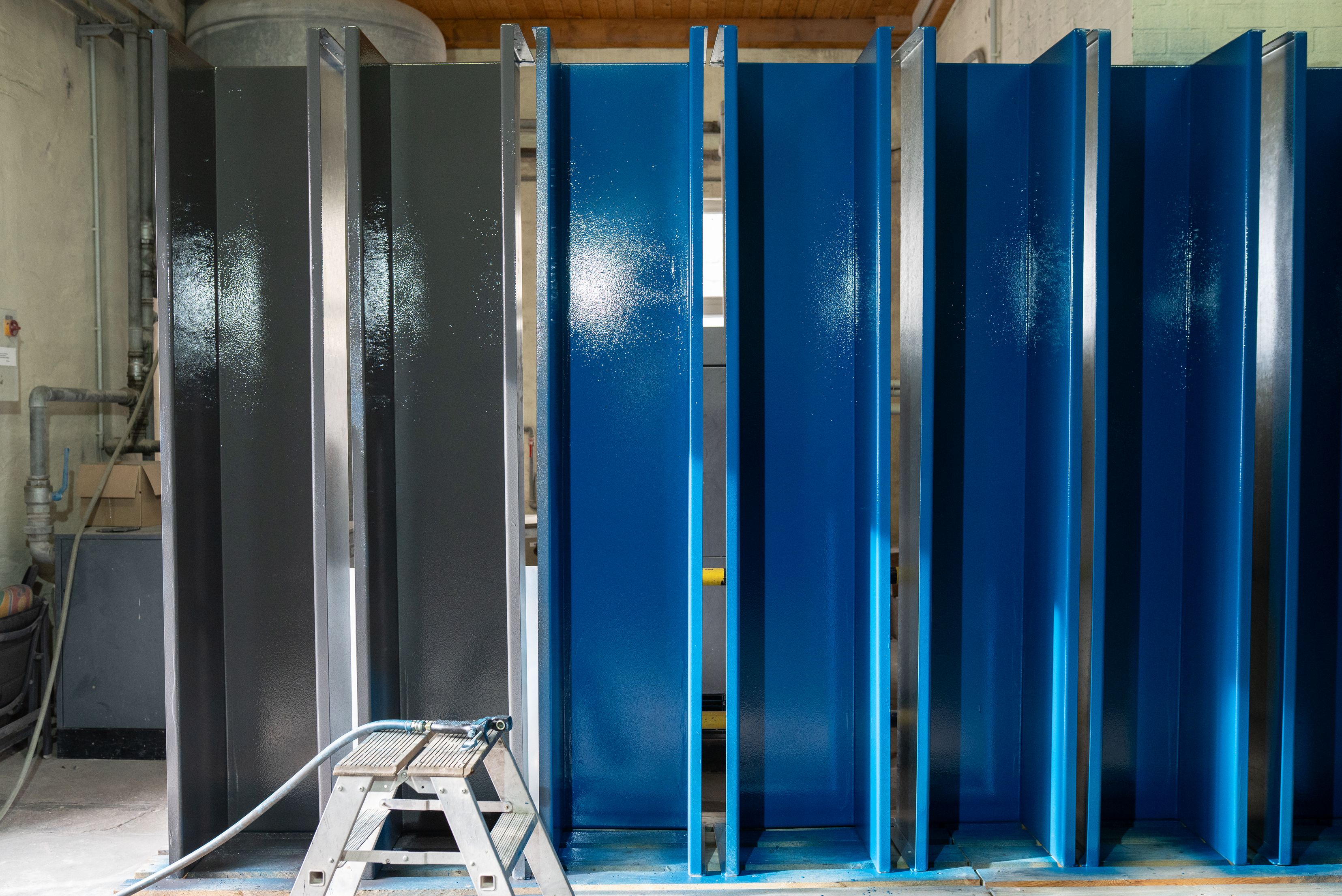A front view of several upright steel beams and metal sections in a workshop, some of which are coated with a grey primer and others with a glossy blue protective coating, with a small folding step ladder in the foreground.