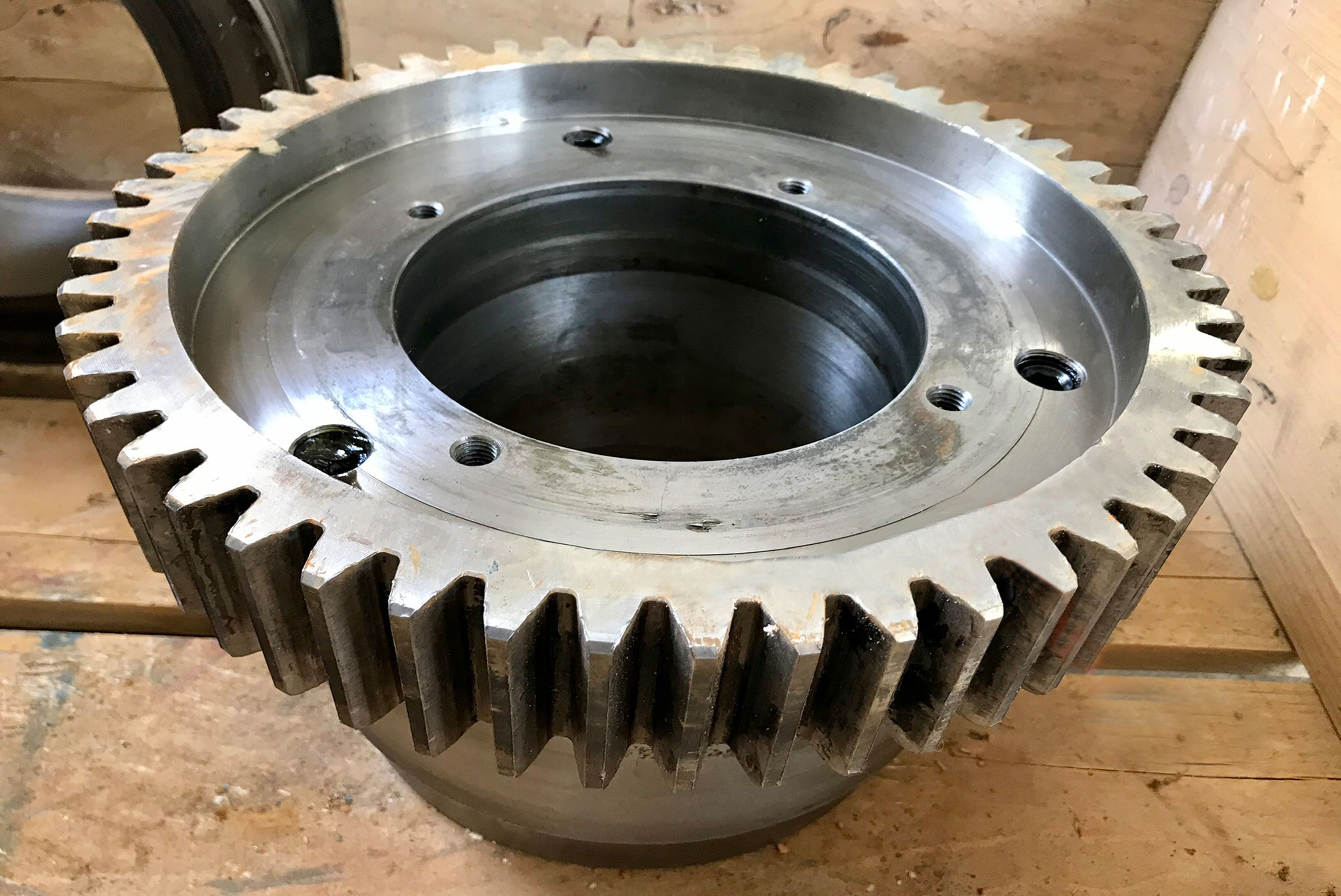 Close-up view of a large, precision-machined steel gear wheel with outer teeth, mounting holes, and a central bore, resting on a wooden pallet after surface cleaning and maintenance.