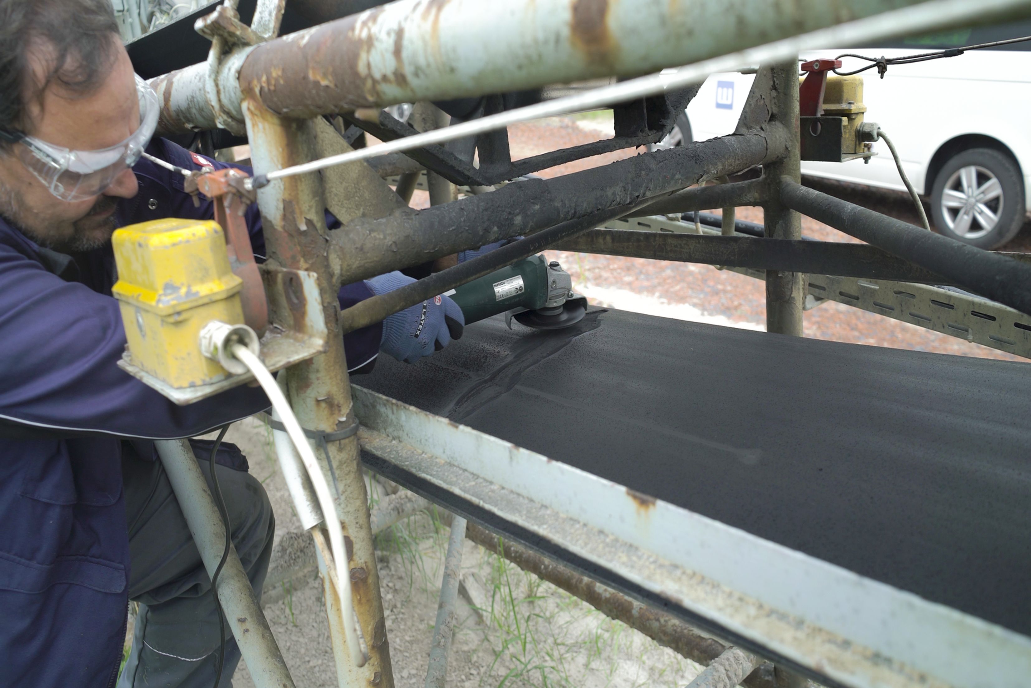 Side view of a technician wearing safety glasses and protective gloves using an angle grinder to prepare or repair the surface of a wide black industrial conveyor belt on an outdoor support frame.