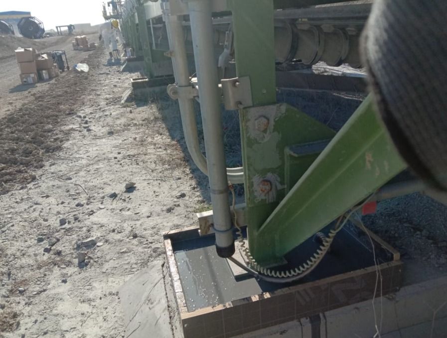 Close-up view of a green-painted steel support foot for an outdoor industrial conveyor belt system, where the concrete foundation has been finished with a dark epoxy casting compound and protective coating.